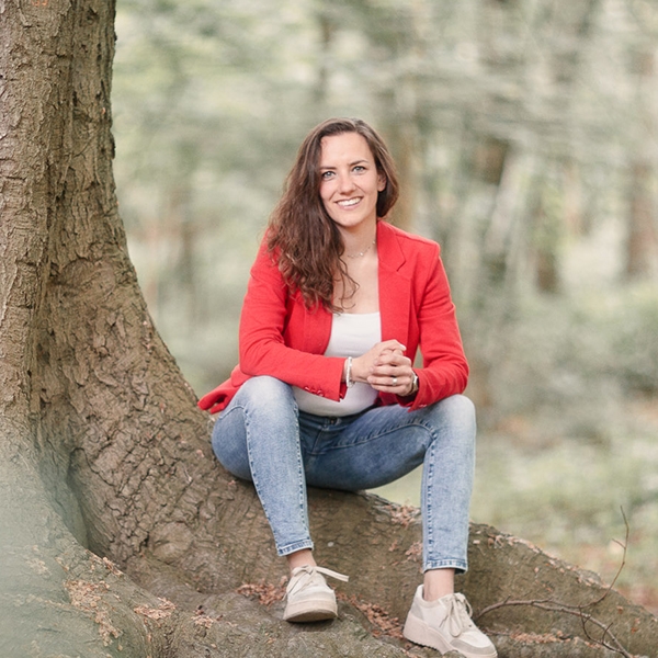 Alicia Zuiderwijk zit buiten op een boomstronk met een grote glimlach, een rood pakjasje en spijkbroek aan.