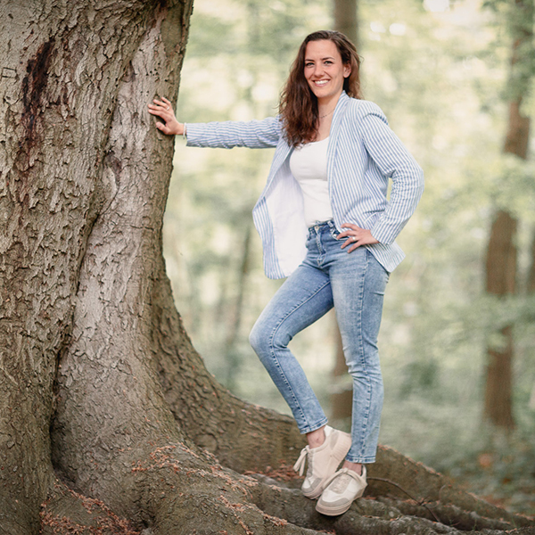 Alicia Zuiderwijk staat tegen een dikke boom aan geleund. Met een grote glimlach met een blauw jasje met witte strepen en een spijkerbroek.